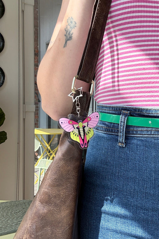 A close-up lifestyle shot of a person wearing a pink and white striped top, blue jeans, and a bright green belt, carrying a large brown leather shoulder bag with a colorful butterfly charm.