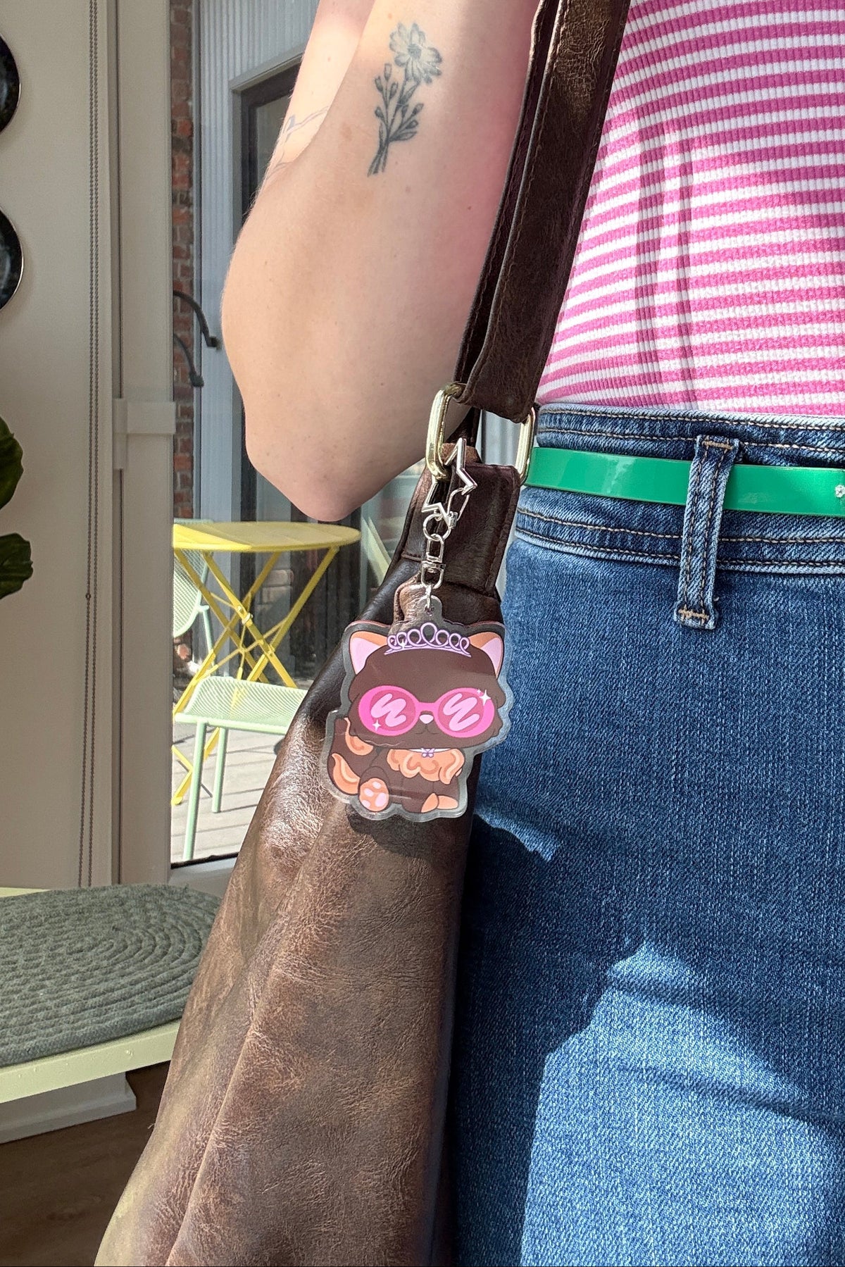 A close-up lifestyle shot of a person wearing a pink and white striped tank top, blue jeans, and a green belt, carrying a brown leather shoulder bag with a pink cat-shaped charm.