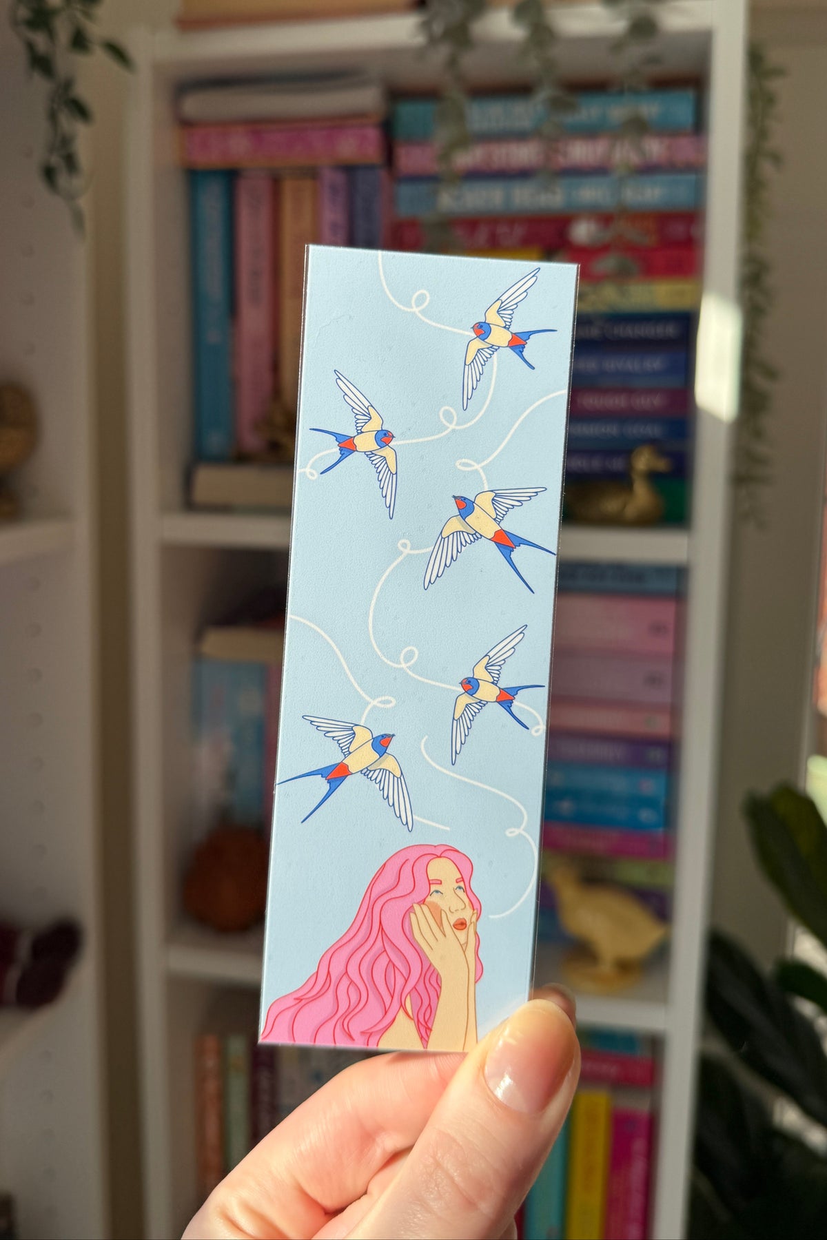 A hand holds a light blue rectangular card or bookmark featuring an illustration of five blue and yellow birds flying in a circle above a pink-haired woman with her hand to her face. The background shows a white bookshelf filled with colorful books in a home setting.