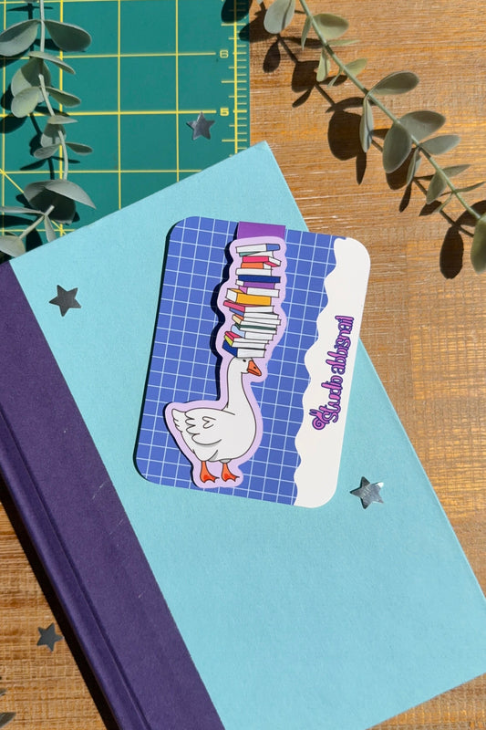 A blue book on a wood table and green cutting mat depicting a magnet bookmark of a goose with a stack of books on its head.