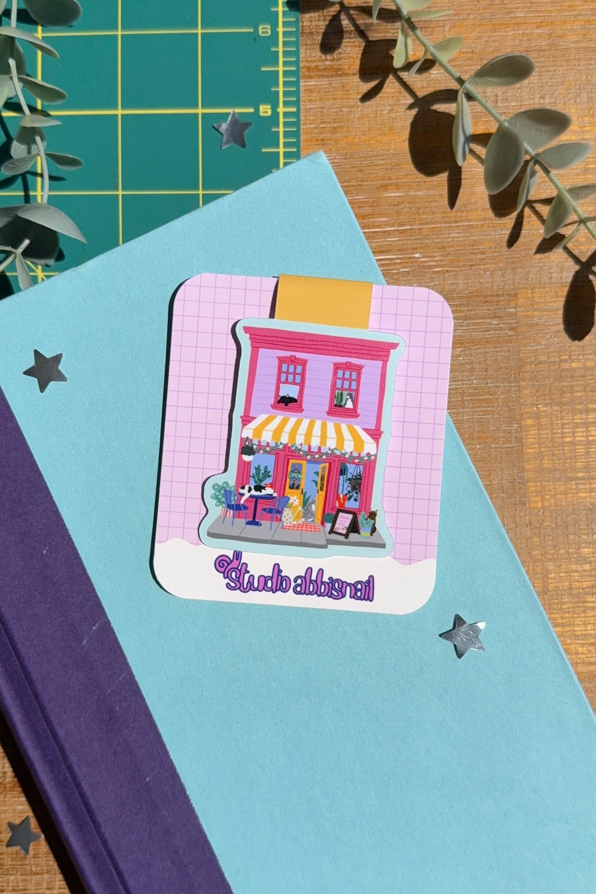 A small illustrated bookmark shaped like a pink storefront with a yellow awning and blue sky is placed on a blue book, surrounded by eucalyptus leaves on a wooden surface with a green cutting mat in the background.