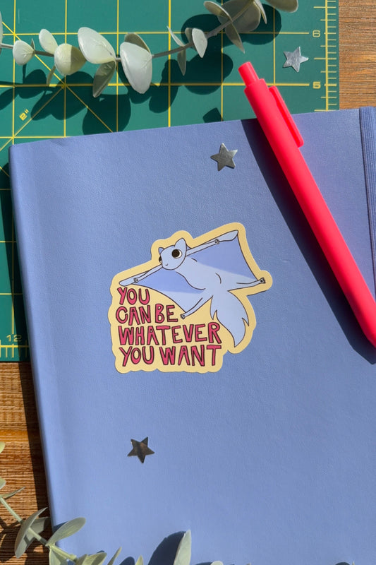 A blue notebook with a die-cut sticker that reads 'YOU CAN BE WHATEVER YOU WANT' with a cartoon flying squirrel, styled on a green cutting mat with a red pen and eucalyptus leaves.