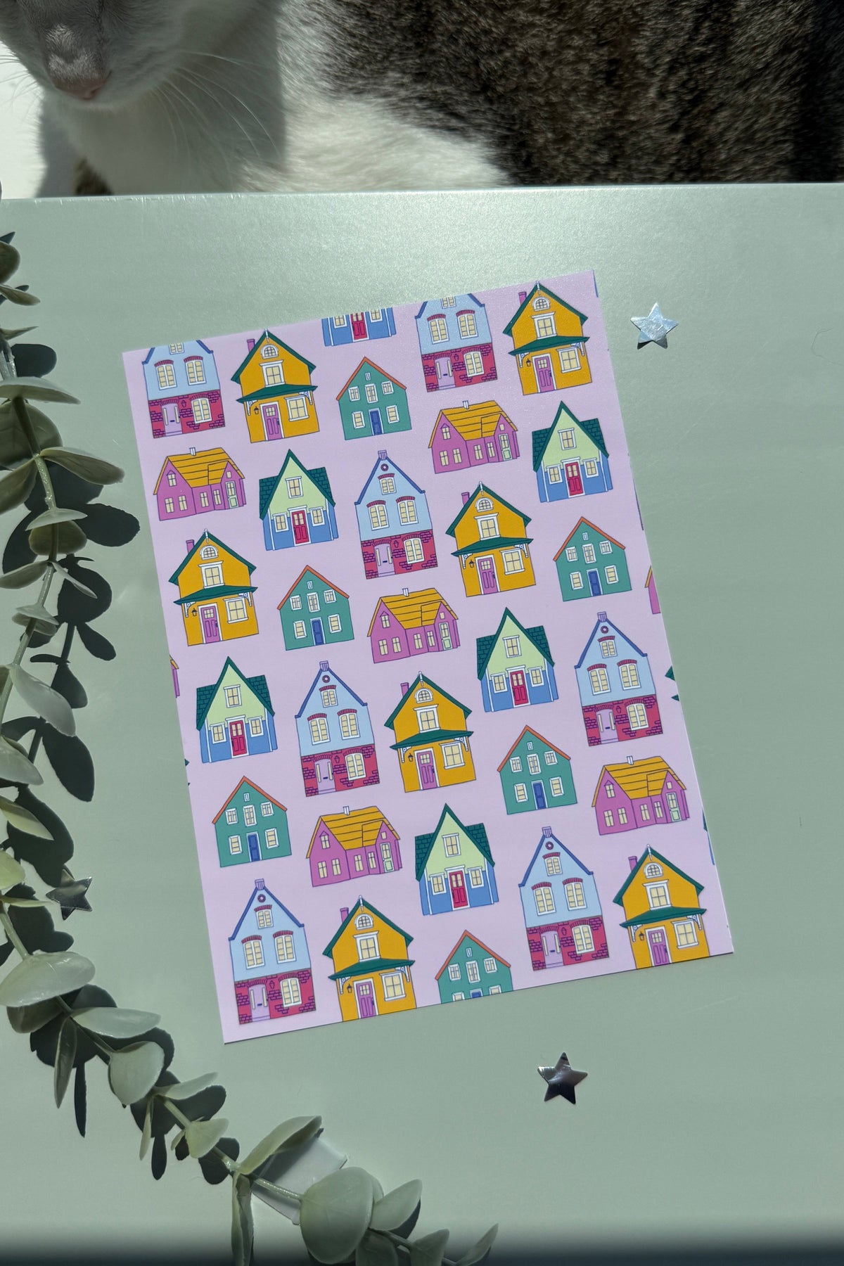 Pink art print featuring colorful whimsical houses on a green surface with a cat visible at the top.