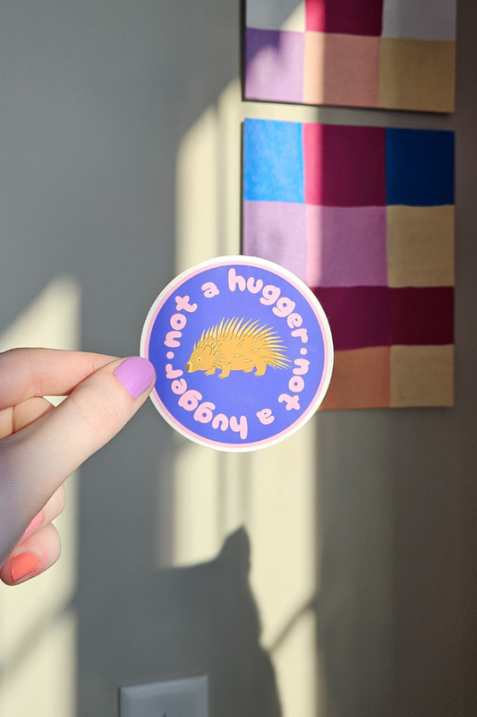 A hand holds a round sticker with a purple background, white text reading 'I'm not a hugger.
