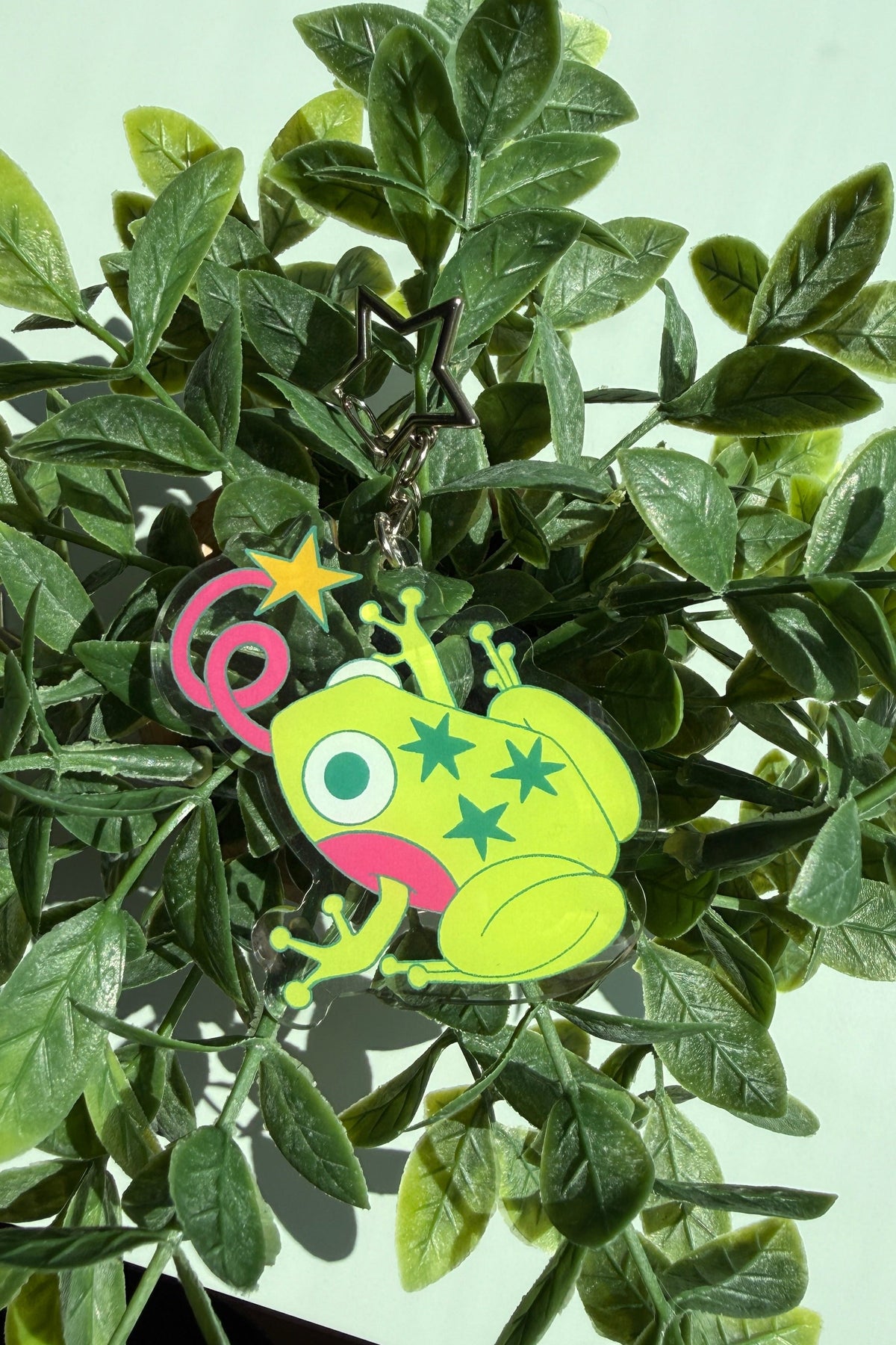 A cartoon frog-shaped keychain charm in bright lime green with pink cheeks, green stars, and a pink swirl tongue, attached to a star keyring and photographed among green leaves.