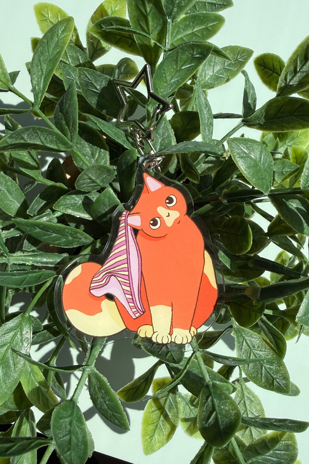 A small illustrated cat-shaped accessory with a pink striped undies, attached to a metal key ring, photographed among green leaves against a pale mint background.