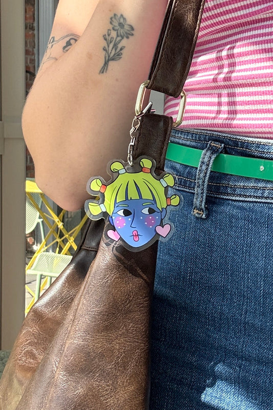 A person wearing a pink and white striped tank top, blue jeans, and a bright green belt, carrying a large brown leather shoulder bag with a colorful cartoon charm.