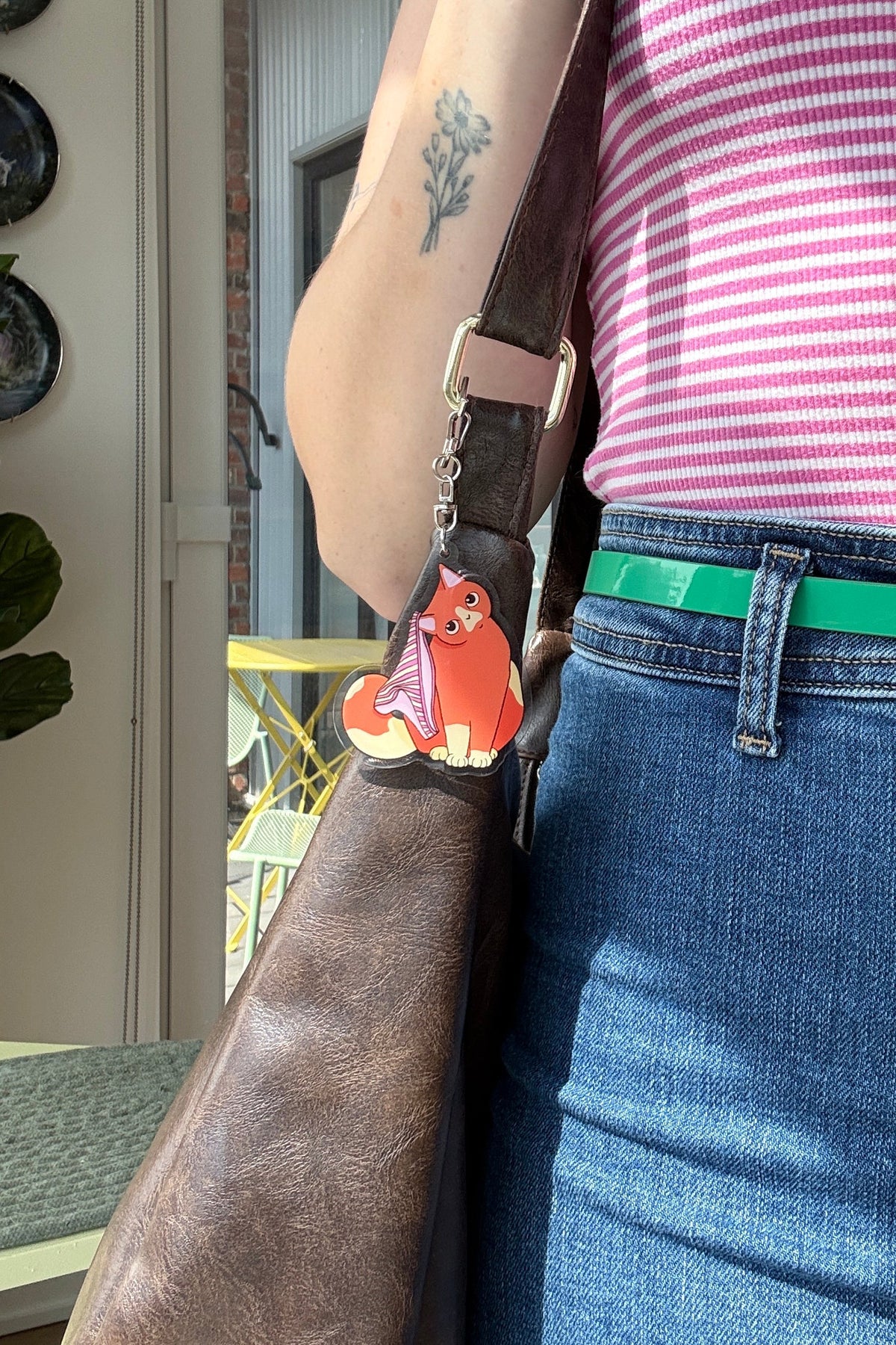 A close-up lifestyle shot of a person wearing a pink and white striped top, blue jeans, and a bright green belt, carrying a brown leather shoulder bag with a small orange cat charm.