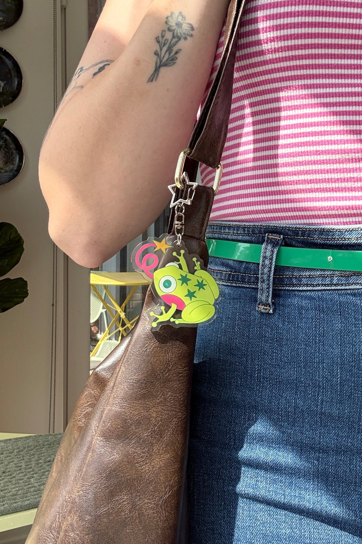 A close-up lifestyle shot of a person wearing a pink and white striped ribbed tank top, blue jeans, and a bright green belt, carrying a brown leather shoulder bag with a colorful frog charm.