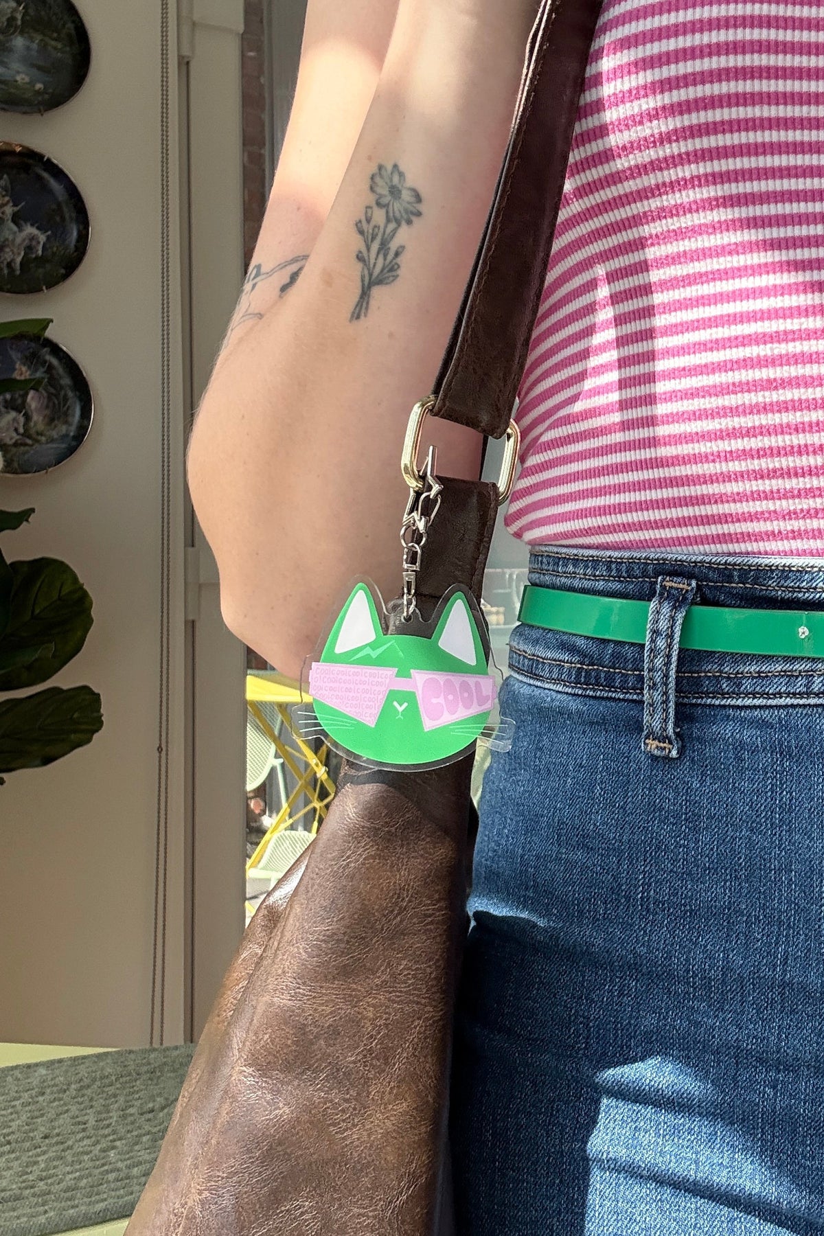A close-up lifestyle shot of a person wearing a pink and white striped tank top, blue jeans, and a bright green belt with a gold buckle. A brown leather shoulder bag is slung over the shoulder, featuring a green and pink cat-shaped charm.