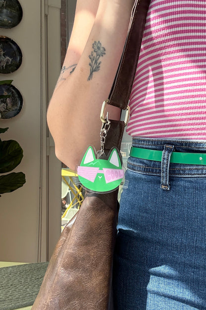 A close-up lifestyle shot of a person wearing a pink and white striped tank top, blue jeans, and a bright green belt with a gold buckle. A brown leather shoulder bag is slung over the shoulder, featuring a green and pink cat-shaped charm.