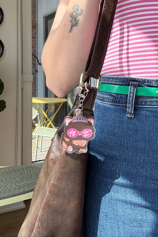A close-up lifestyle shot of a person wearing a pink and white striped tank top, blue jeans, and a green belt, carrying a brown leather shoulder bag with a pink cat-shaped charm.