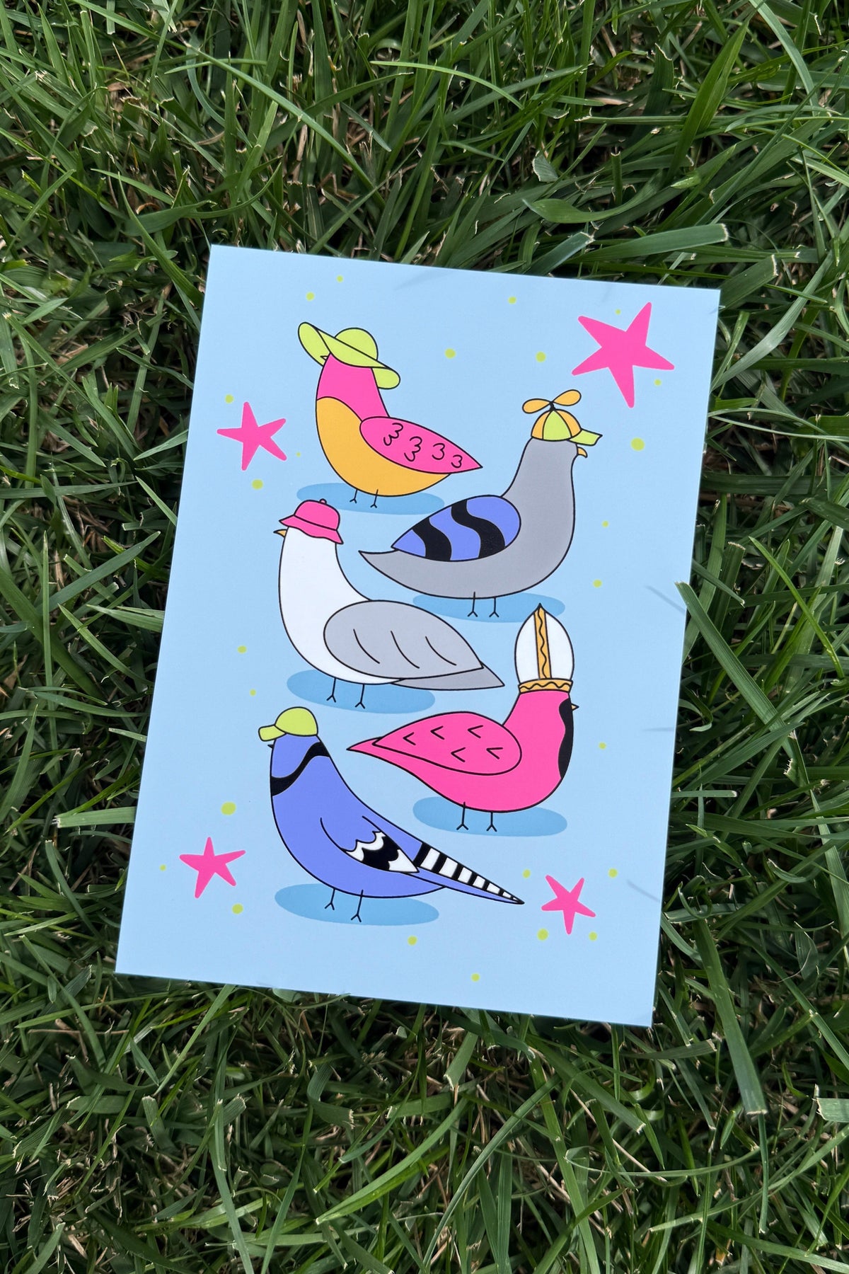 A square art print featuring a whimsical illustration of five birds in pink, yellow, blue, gray, and white on a light blue background, photographed lying on green grass.