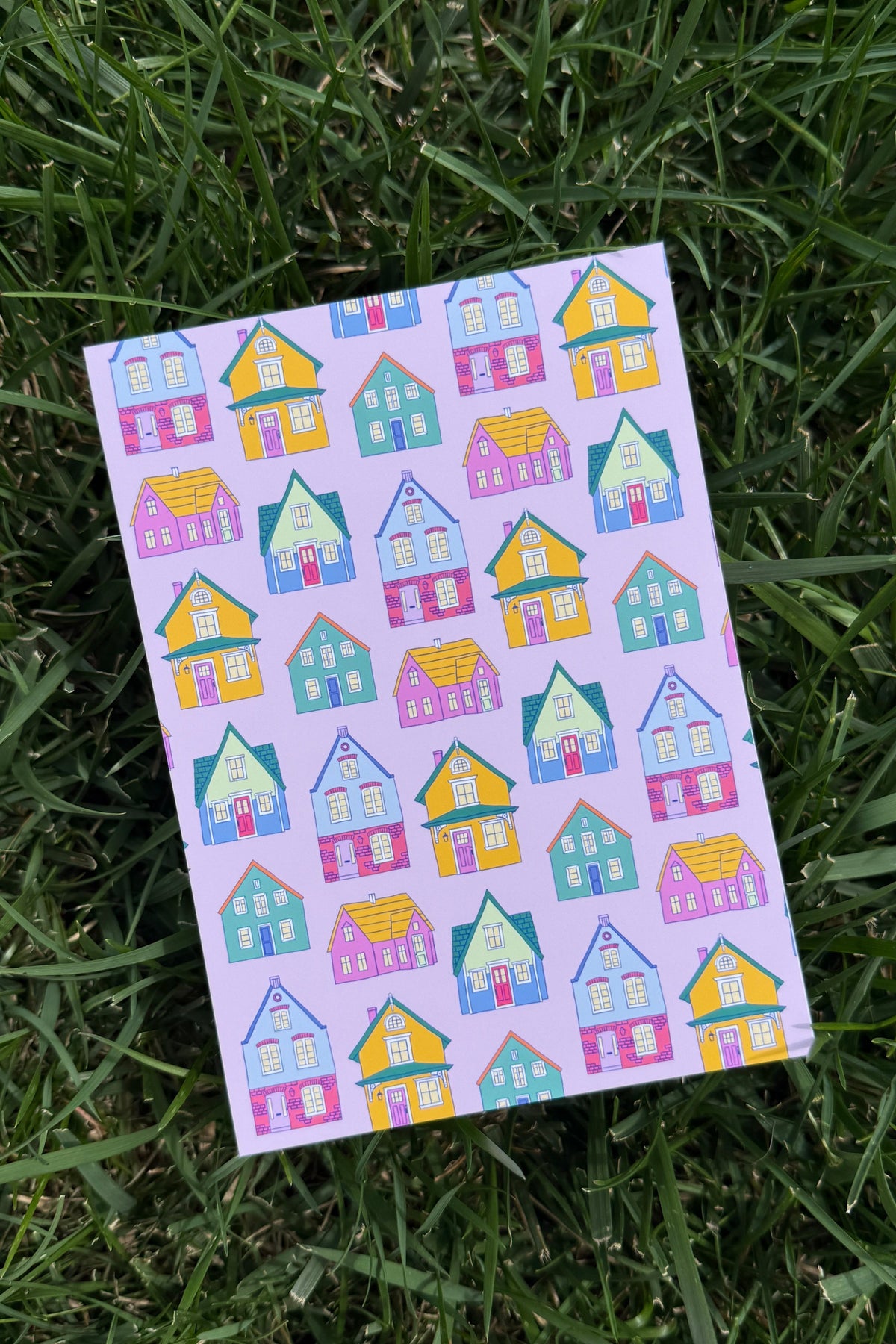 A rectangular art print with a repeating pattern of colorful cartoon houses, placed on green grass.