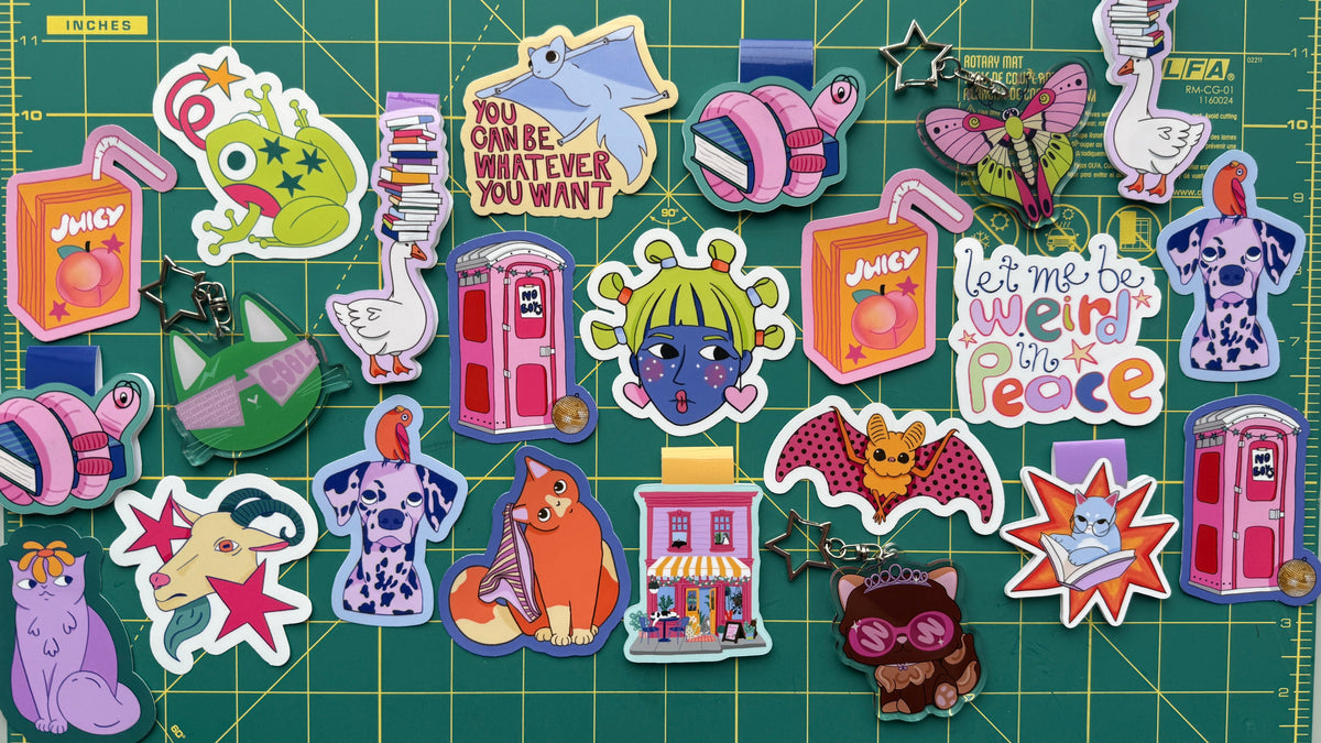 A collection of colorful cartoon and pop culture enamel pins arranged on a green cutting mat with measurement markings. The pins feature characters and designs inspired by The Simpsons, including Bart, Lisa, Maggie, Homer, and Marge, alongside pop culture references like the Joker, Spider-Man, and Star Wars.