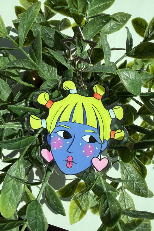 A colorful cartoon face keychain with a blue face, yellow hair, and pink hearts, hanging from a metal star clip among green leaves.