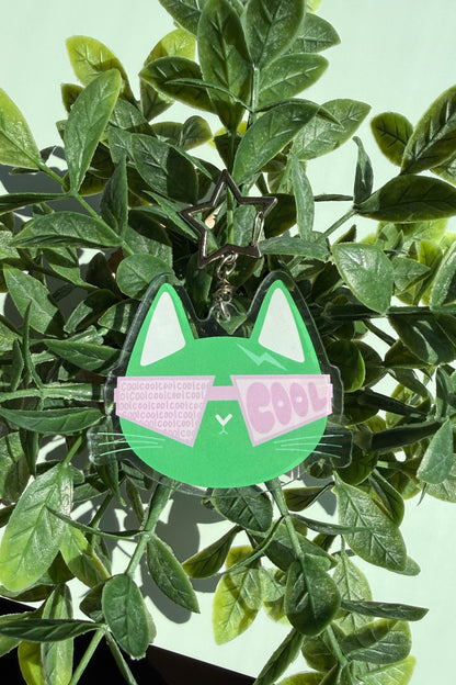 A green cat-shaped acrylic keychain with pink sunglasses and the word 'COOL' printed on them, displayed among green leaves.