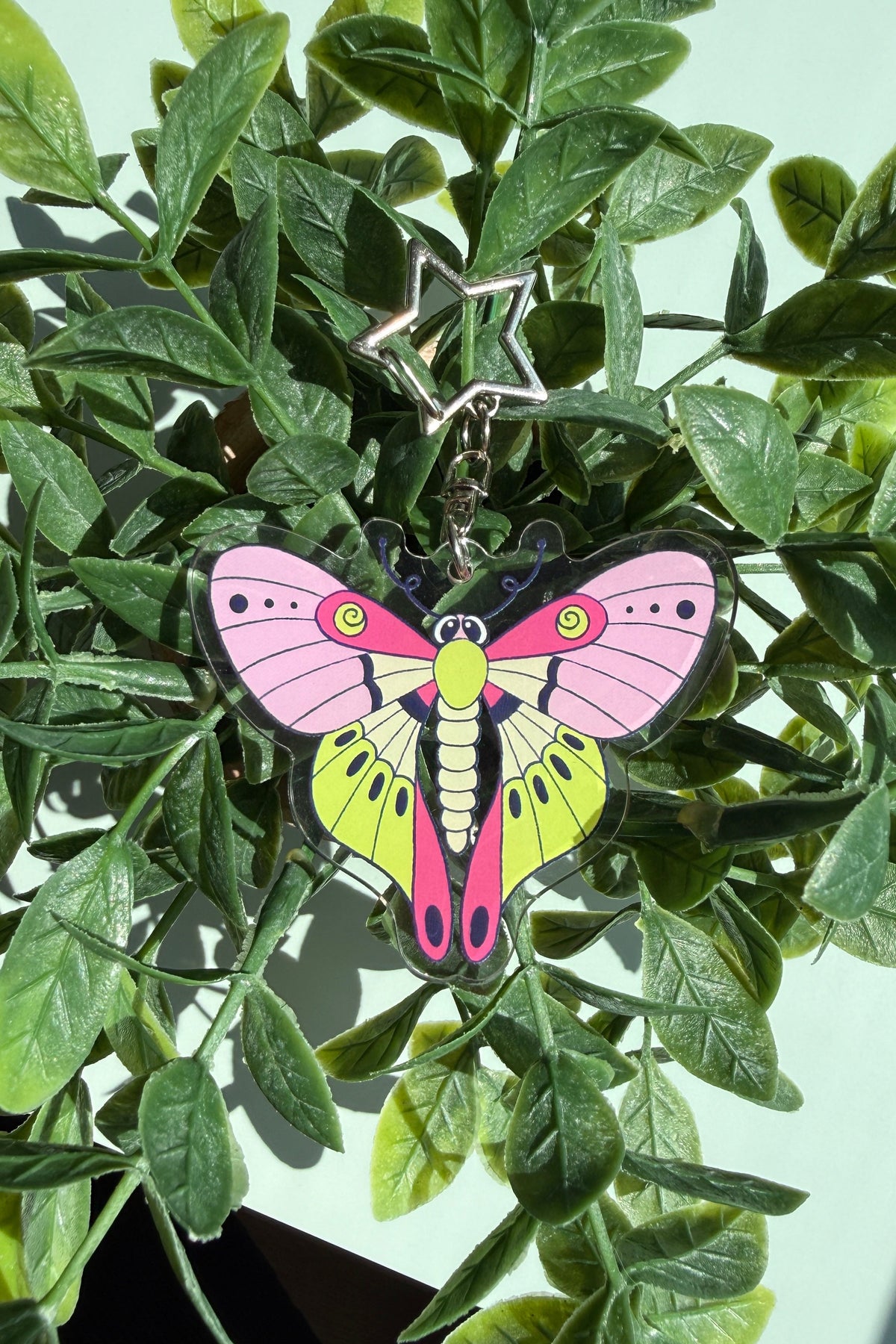 A colorful butterfly-shaped keychain with pink, yellow, and green wings hanging from a silver star key ring, displayed among green artificial leaves.