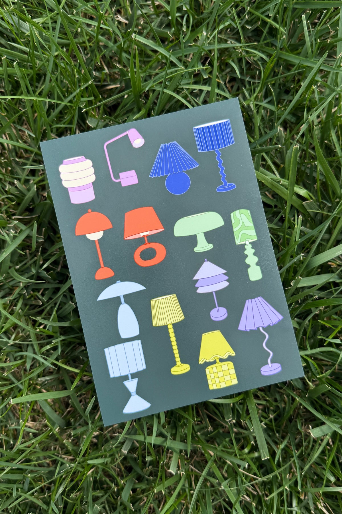 A rectangular art print featuring a collection of colorful, retro-style lamp illustrations on a dark green background, photographed lying on green grass.