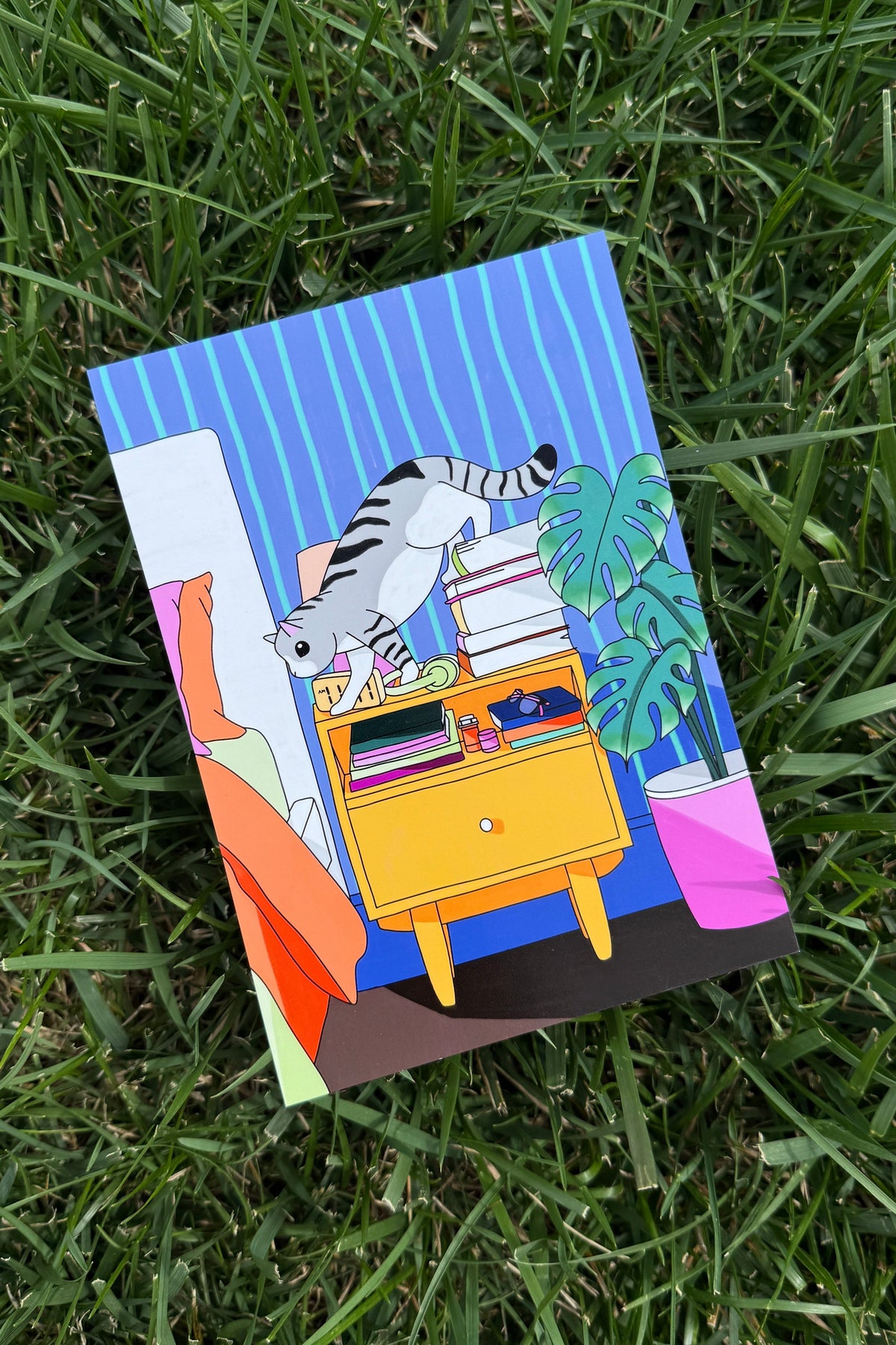 A colorful art print depicting a cartoon cat climbing on books next to a yellow cabinet, placed on a green grass background.