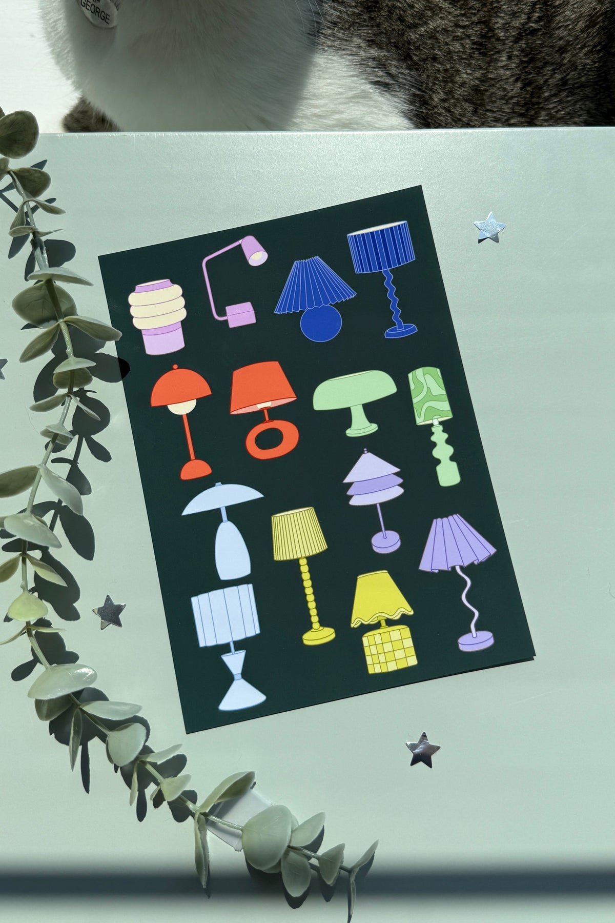 A square art print featuring a colorful collection of lamp illustrations on a dark background, styled on a light surface with eucalyptus leaves and star stickers, with a cat partially visible at the top.