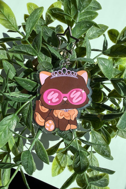 A cute cartoon keychain featuring a brown cat wearing pink sunglasses and a tiara, hanging among green leaves.