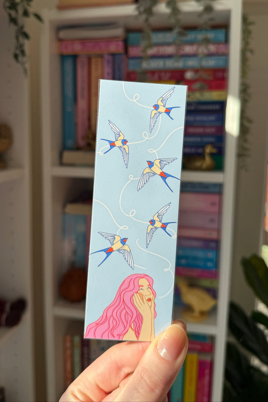 A hand holds a light blue rectangular card or bookmark featuring an illustration of five blue and yellow birds flying in a circle above a pink-haired woman with her hand to her face. The background shows a white bookshelf filled with colorful books in a home setting.