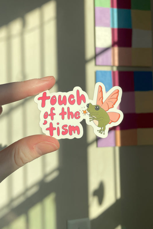 A hand holds a die-cut sticker that reads 'touch of the 'tism' in pink lettering next to an illustration of a green frog with pink wings. The background shows a wall with colorful square panels and window shadow light.