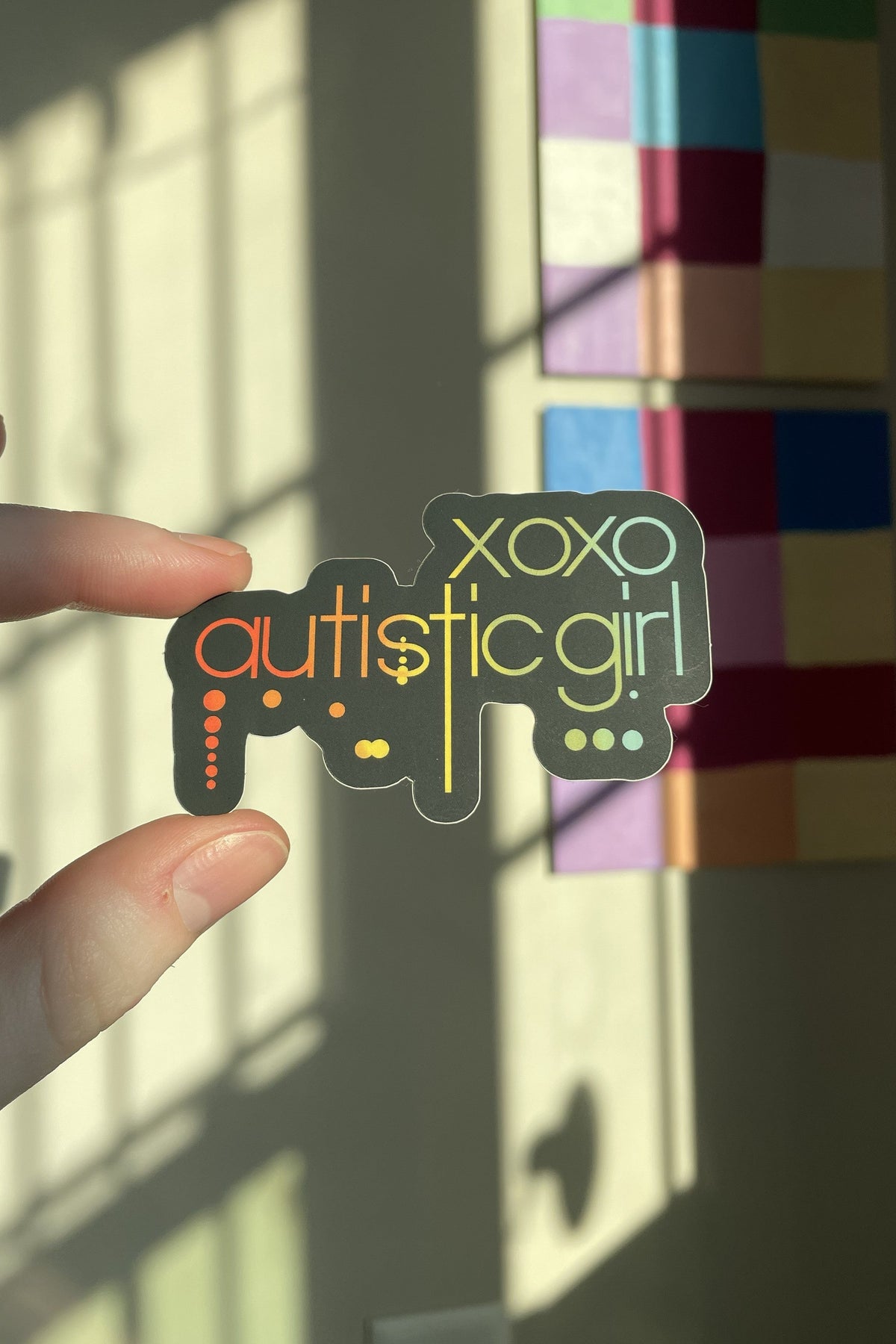 A hand holds a die-cut sticker that reads 'xoxo autistic girl' with a controller graphic, set against a wall with colorful art panels and window shadows.