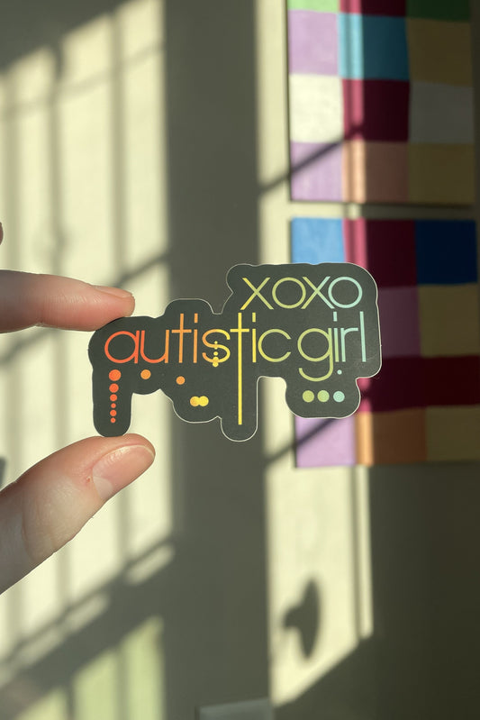 A hand holds a die-cut sticker that reads 'xoxo autistic girl' with a controller graphic, set against a wall with colorful art panels and window shadows.