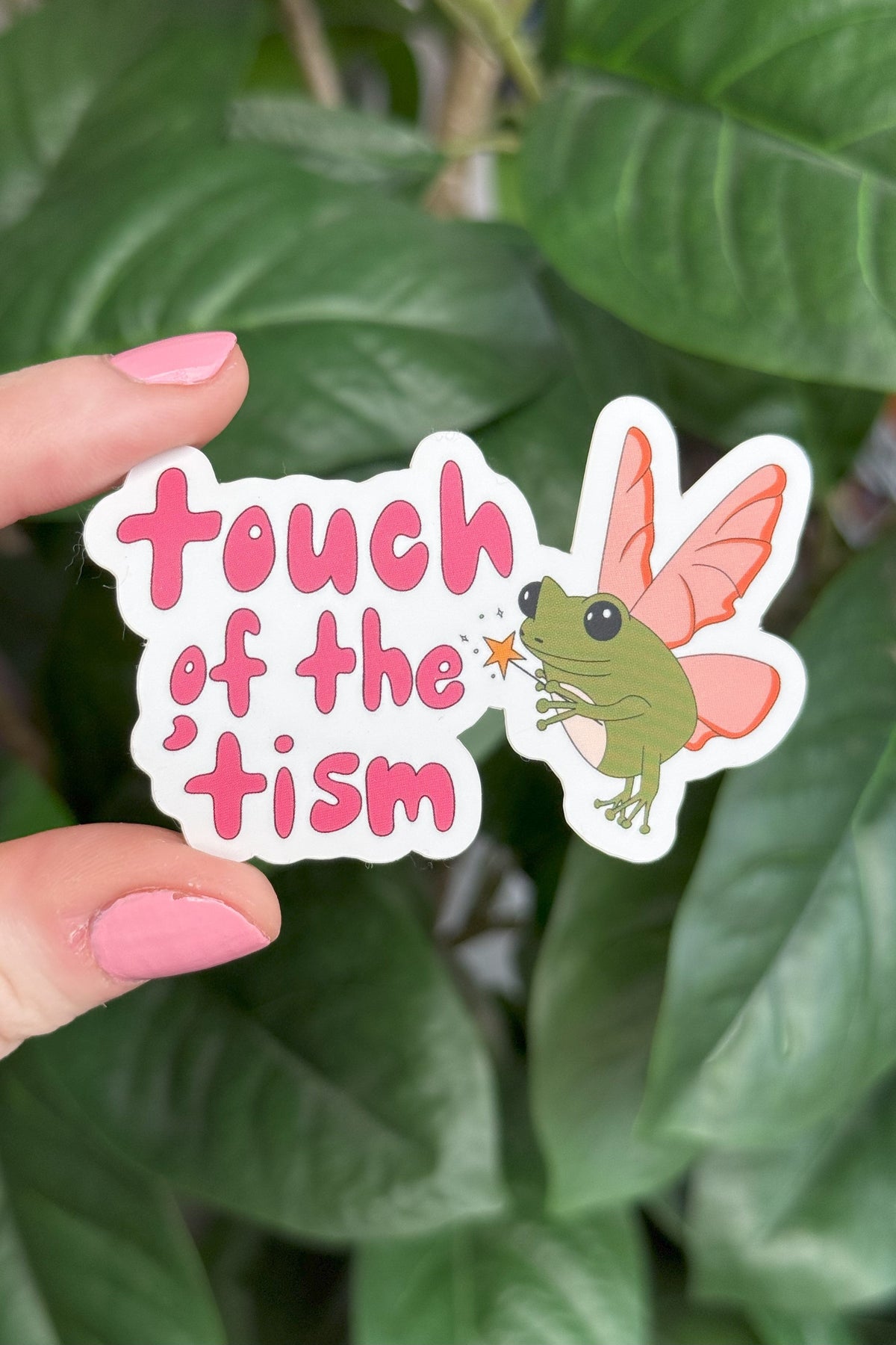 A die-cut sticker held by a hand with pink nail polish against a background of green houseplant leaves. The sticker features the text 'touch of the 'tism' in pink lettering next to an illustration of a green fairy with pink wings.