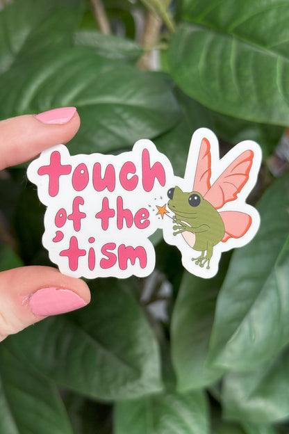A die-cut sticker held by a hand with pink nail polish against a background of green houseplant leaves. The sticker features the text 'touch of the 'tism' in pink lettering next to an illustration of a green fairy with pink wings.