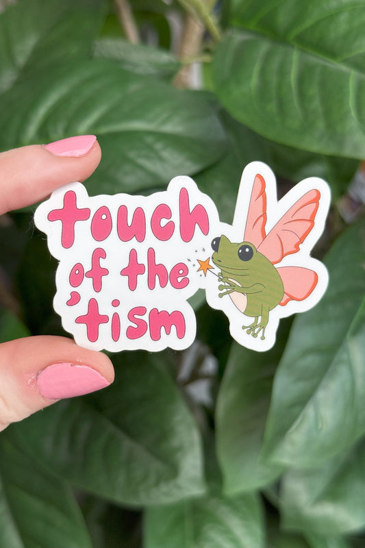 A die-cut sticker held by a hand with pink nail polish against a background of green houseplant leaves. The sticker features the text 'touch of the 'tism' in pink lettering next to an illustration of a green fairy with pink wings.