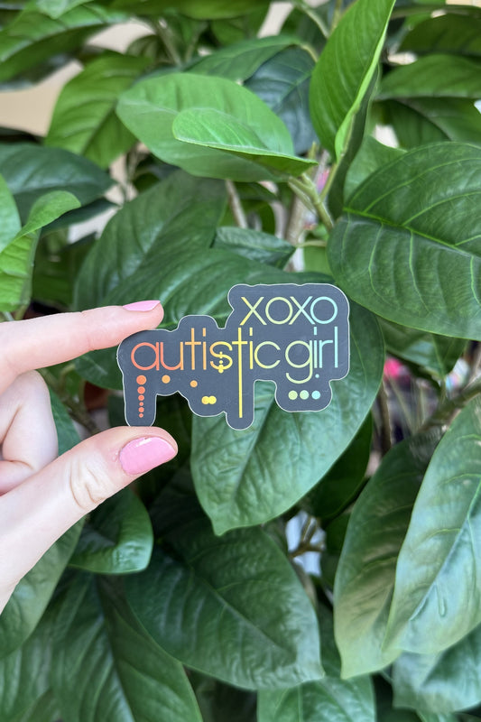 A hand with pink nail polish holds a small die-cut sticker in front of a green leafy houseplant. The sticker reads 'xoxo autistic girl' in stylized lettering.