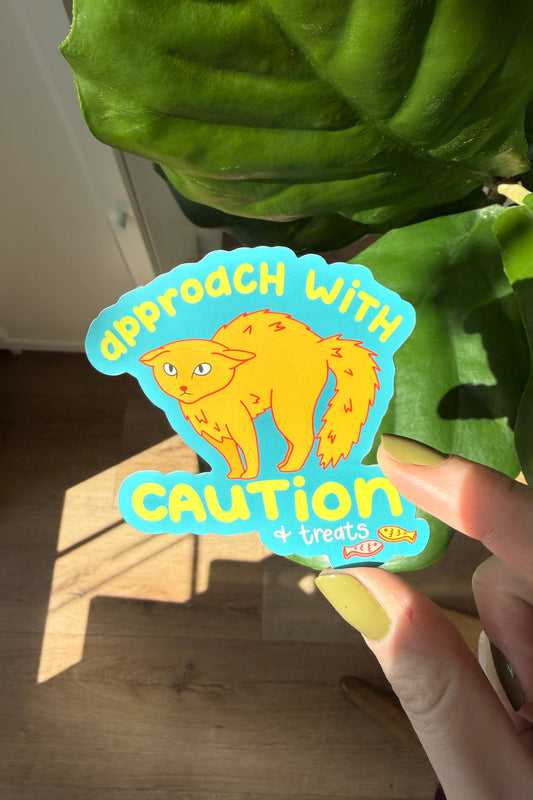 A hand holds a die-cut sticker featuring a cartoon orange cat and the text 'approach with CAUTION + treats only' against a background of a green plant and wooden floor.