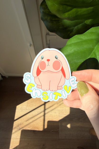 A hand holds a die-cut sticker featuring a cute pink bunny and the word 'SENSITIVE' in colorful bubble letters, with a real indoor environment, wooden floor, and green houseplant in the background.