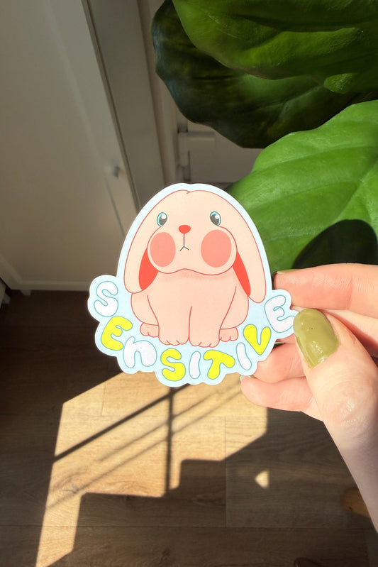 A hand holds a die-cut sticker featuring a cute pink bunny and the word 'SENSITIVE' in colorful bubble letters, with a real indoor environment, wooden floor, and green houseplant in the background.