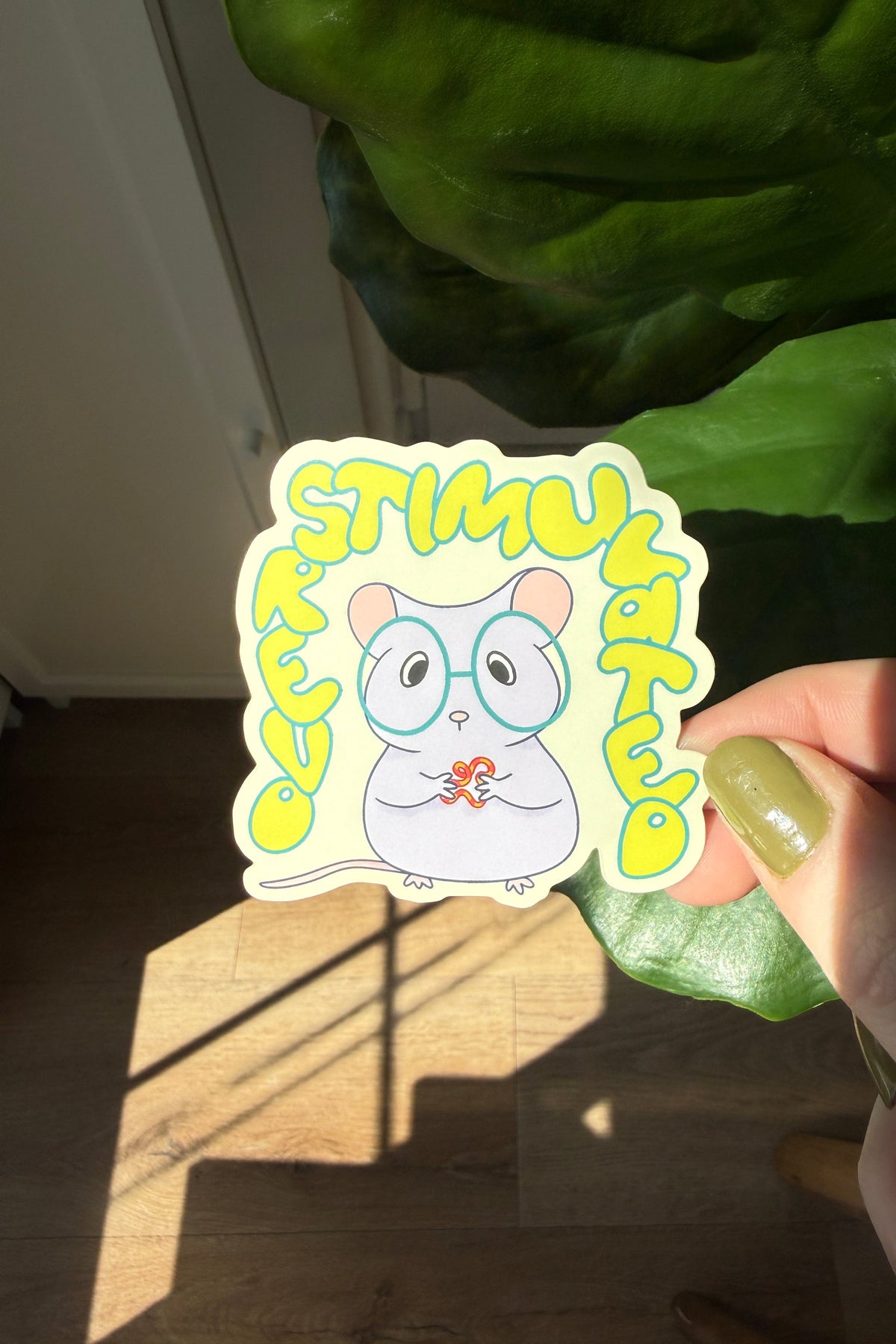 A hand with green nail polish holds a die-cut sticker featuring a cartoon mouse wearing glasses and the text 'OVERSTIMULATED' in yellow, set against a background of a wooden floor, a plant, and warm sunlight.