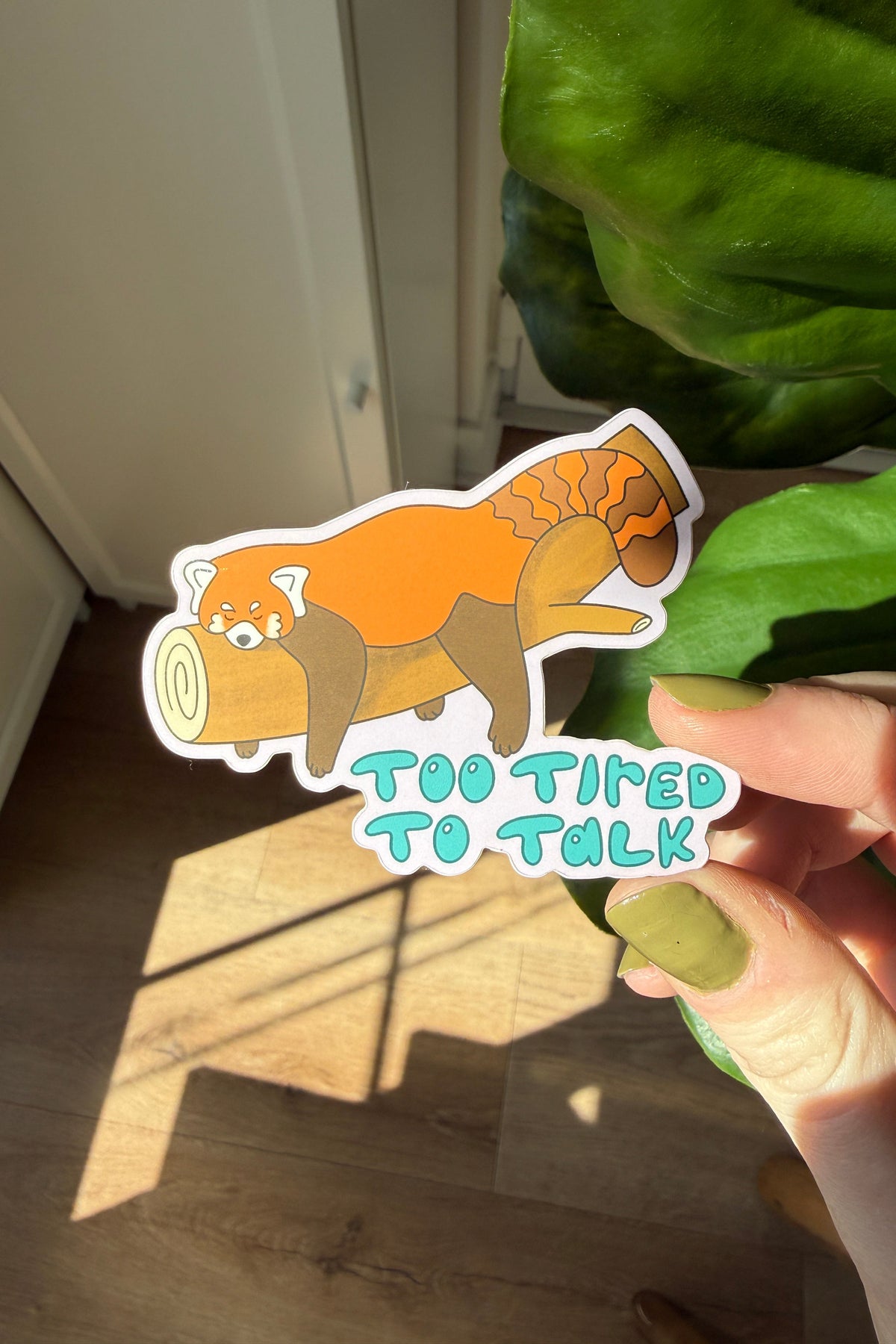 A die-cut sticker featuring a cartoon red panda sitting on a log with the text 'TOO TIRED TO TALK' in light blue letters, held by a hand in front of a green plant and warm sunlight.
