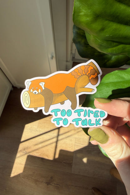 A die-cut sticker featuring a cartoon red panda sitting on a log with the text 'TOO TIRED TO TALK' in light blue letters, held by a hand in front of a green plant and warm sunlight.