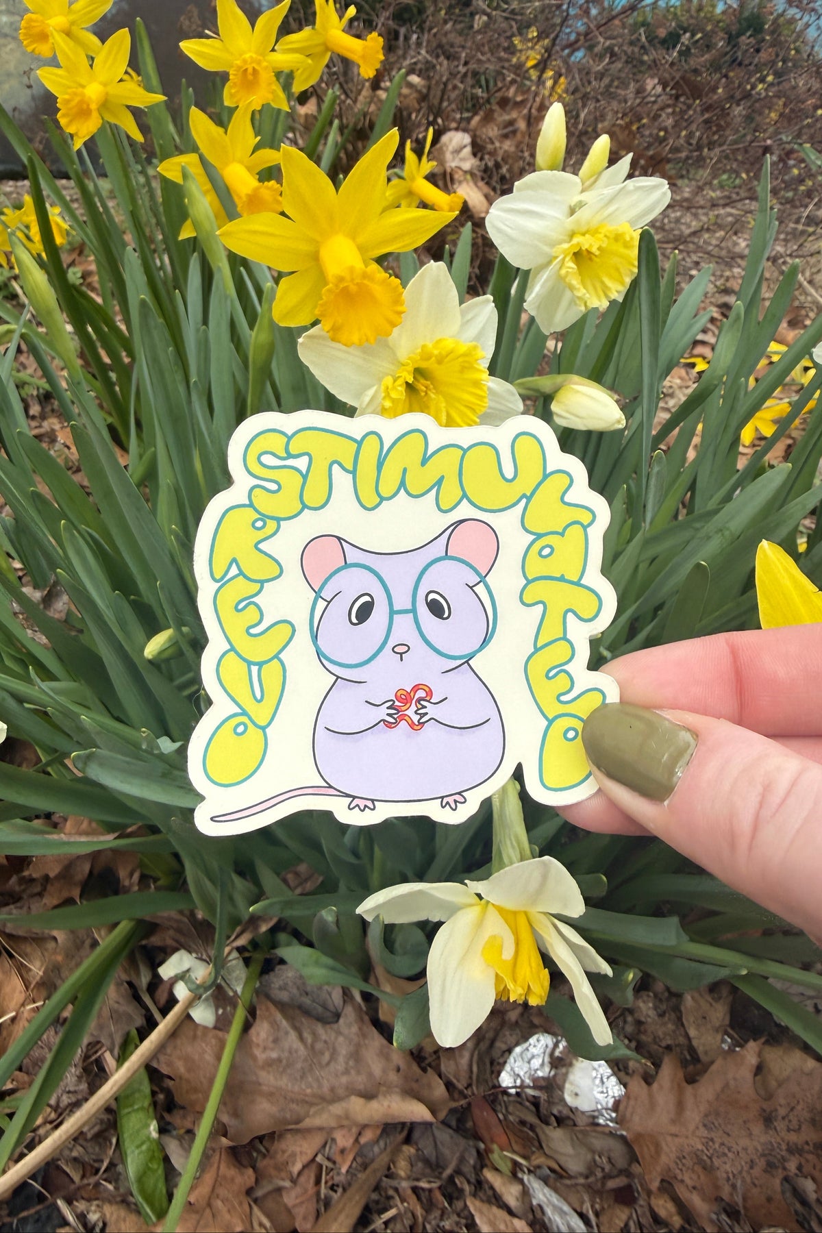 A hand holds a die-cut sticker featuring a cute lavender cartoon mouse wearing round glasses and holding a small red flower, surrounded by yellow daffodils and green leaves outdoors.