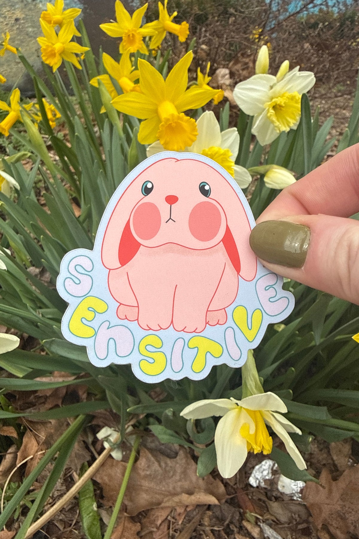 Die cut sticker of a pink bunny and the word "SENSITIVE" being held in front of yellow daffodils