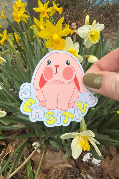 Die cut sticker of a pink bunny and the word "SENSITIVE" being held in front of yellow daffodils