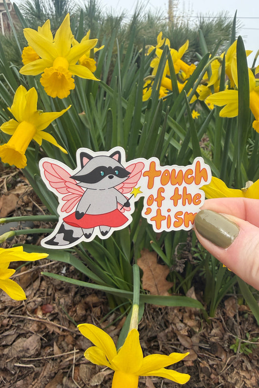A hand with olive green nail polish holds a die-cut sticker featuring a cartoon raccoon fairy with pink wings and a red dress next to the text 'touch of the tism'. The background is a garden filled with blooming yellow daffodils.