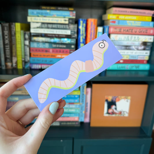 A hand holding a lavender bookmark featuring a cartoon worm illustration, with a blurred bookshelf in the background.
