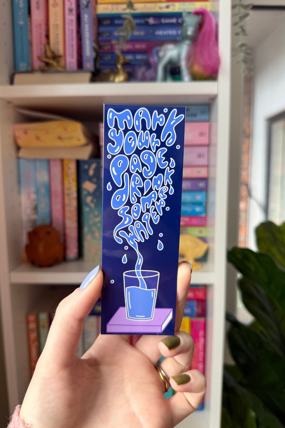 A hand holds a dark blue rectangular card or coaster featuring a cartoon glass of blue liquid with the text 'mark your page, drink some water' in a bubbly light blue font. The background shows a blurred bookshelf with colorful books and a plant.