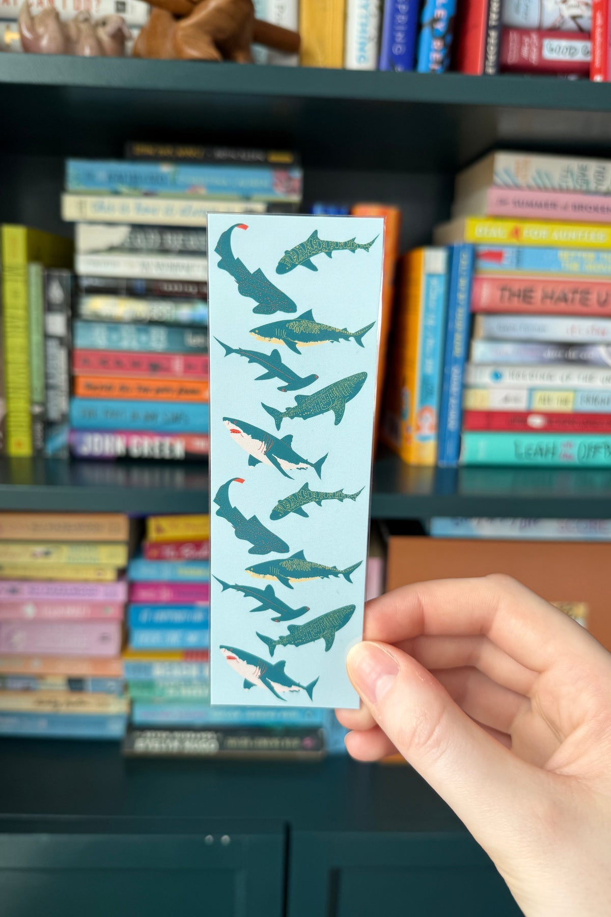 A hand holds a long, narrow light blue bookmark or card featuring a repeating pattern of dark teal sharks, with small red accents, in front of a blurred bookshelf.