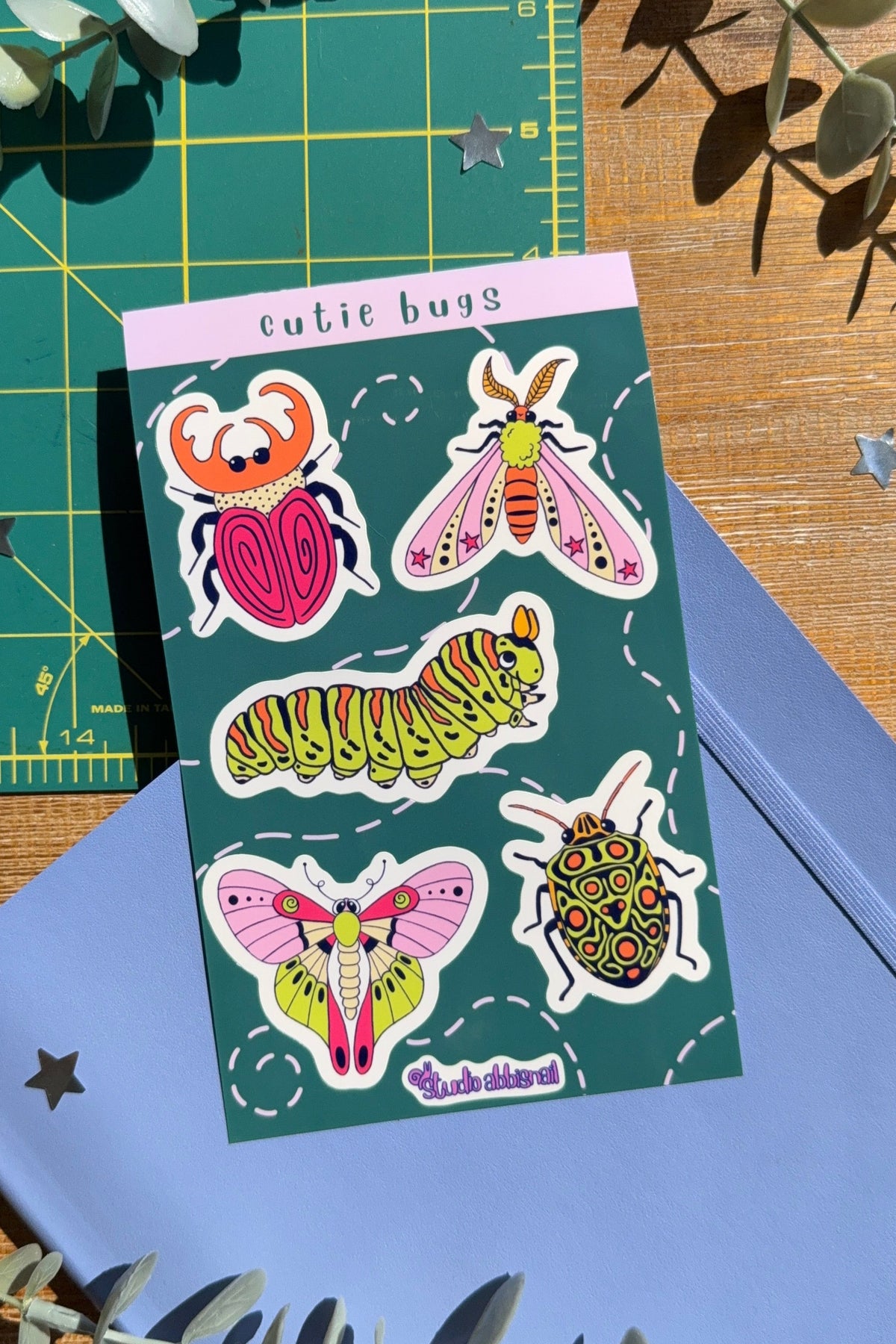 Sticker sheet titled 'cutie bugs' with drawings of bugs on a blue notebook with a wood desk background.