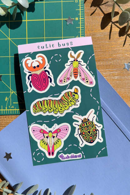 Sticker sheet titled 'cutie bugs' with drawings of bugs on a blue notebook with a wood desk background.