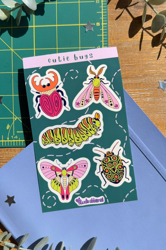 Sticker sheet titled 'cutie bugs' with drawings of bugs on a blue notebook with a wood desk background.